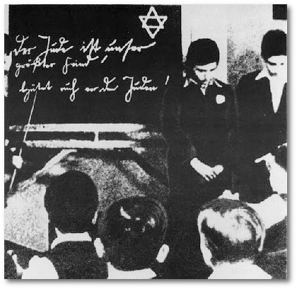 Um 1935: Zwei jüdische Schüler werden vor ihren Klassenkameraden gedemütigt. An der Tafel antisemitische Hetze: 'Der Jude ist unser größter Feind! Schützt Euch vor den Juden!' 